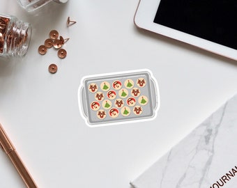 May include: A silver baking sheet filled with Christmas-themed sugar cookies. The cookies are decorated with red and green icing and feature images of Santa Claus, reindeer, and Christmas trees.