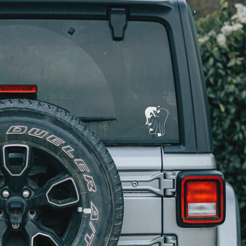 May include: White vinyl car decal featuring a ghost embracing a cat. The decal is affixed to the rear window of a silver SUV, with a black spare tire and a red taillight visible.