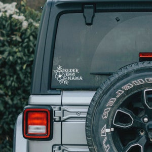 May include: A silver Jeep with a white vinyl sticker on the back window. The sticker features a floral design and the text "Bloomer Emo Mama".