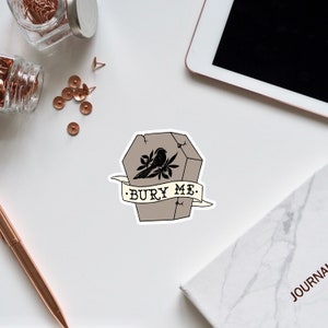 May include: A sticker with a black and white illustration of a coffin with a bird perched on top. The coffin is on a beige background and the text "BURY ME." is written in black on a white banner.