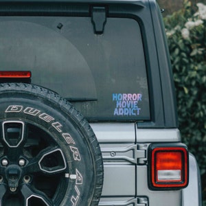 May include: A silver Jeep Wrangler with a black spare tire and a sticker on the back window that reads "HORROR MOVIE ADDICT" in a rainbow gradient.