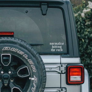 May include: A silver Jeep with a black spare tire and a white vinyl sticker on the back window that reads "normalize hitting the curb".
