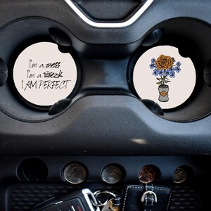 May include: Two white round car coasters. One coaster has the text "I'm a mess, I'm a wreck, I AM PERFECT". The other coaster has a drawing of a bouquet of flowers in a can.