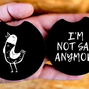 May include: Two black round coasters with white designs. One coaster features a cartoon bird with a white body and black wings. The other coaster has the text "I'm not sad anymore" in white.