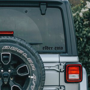 May include: A silver car with a black tire and a black sticker on the back window that says "elder emo".