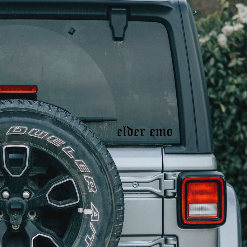 May include: A silver car with a black tire and a black sticker on the back window that says "elder emo".