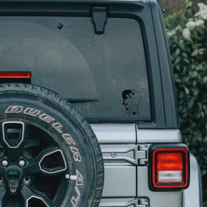 May include: Black vinyl car decal featuring a ghost with a cat on its head. The decal is affixed to the rear window of a silver SUV. The spare tire is visible on the left side of the image.