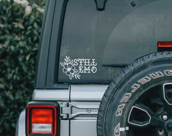May include: A silver car with a black tire and a white sticker on the back window that says "Still Emo" with a floral design.