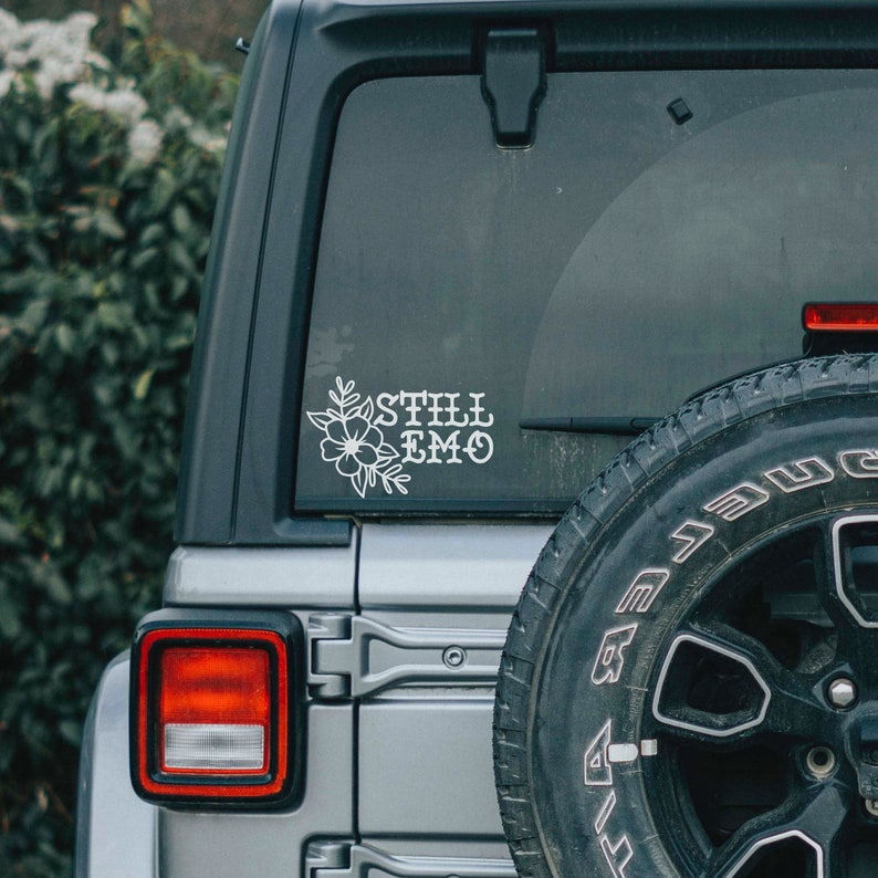 Puede incluir: Un coche plateado con una llanta negra y una pegatina blanca en la ventana trasera que dice "Still Emo" con un dise&ntilde;o floral.