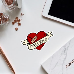 May include: A red heart sticker with a bandage and the words "Still Emo" on a banner. The heart has stitched lines and a white border. The sticker is on a white surface with office supplies.