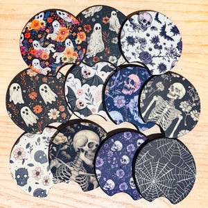 May include: A collection of Halloween-themed car coasters. The coasters feature various designs, including ghosts, skulls, bats, flowers, and spiderwebs. The color palette includes black, white, purple, and orange. The coasters are round with a small notch.