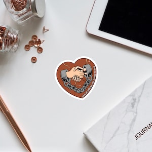 May include: A heart-shaped sticker with a cracked brown background. Two hands are shown shaking, each hand is shackled with a chain. The text reads "Don't be looking for a hand to hold".
