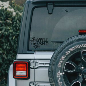 Puede incluir: Un coche plateado con una pegatina floral negra que dice "Still Emo" en la ventana trasera.