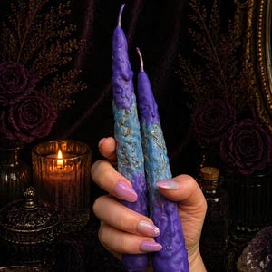 May include: Two tapered candles featuring a textured, uneven surface with a color gradient transitioning from deep purple at the base to a lighter blue in the middle, accented with gold leaf flakes throughout the midsection and a purple top.