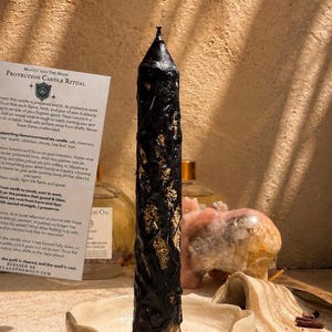 May include: A tall, black, textured pillar candle embedded with gold-colored flakes, standing upright in a shallow, off-white ceramic dish. Beside the candle is a white paper card titled Protection Candle Ritual with printed black text.