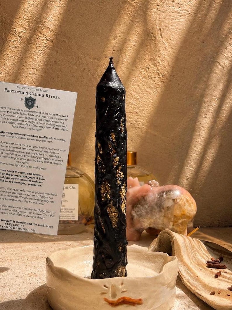 May include: A tall, black, textured pillar candle embedded with gold-colored flakes, standing upright in a shallow, off-white ceramic dish. Beside the candle is a white paper card titled Protection Candle Ritual with printed black text.