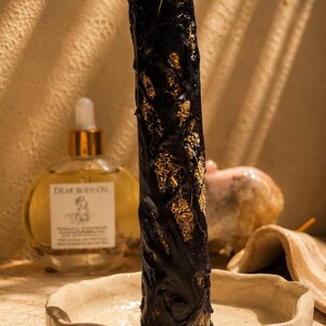 May include: A tall, cylindrical black candle with a textured, vine-like exterior revealing gold-colored flecks, standing upright in a shallow, off-white ceramic dish. Behind it sits a round glass bottle of Dear Body Oil with a gold dropper cap.