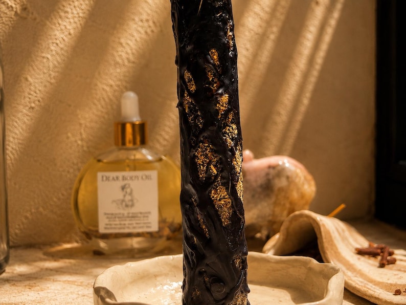 May include: A tall, cylindrical black candle with a textured, vine-like exterior revealing gold-colored flecks, standing upright in a shallow, off-white ceramic dish. Behind it sits a round glass bottle of Dear Body Oil with a gold dropper cap.