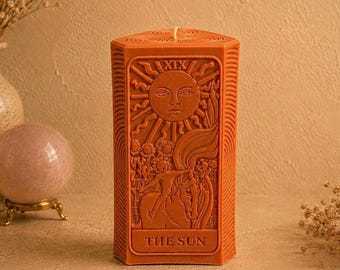 May include: A rectangular terracotta-colored wax candle featuring a relief design of The Sun tarot card. The front displays a large sun face, a child riding a horse, sunflowers, and the text XIX and THE SUN. The candle has a single wick.