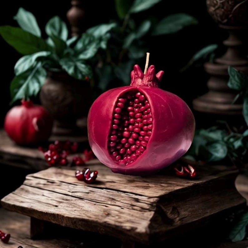 May include: A red pomegranate-shaped candle with a detailed, open-section design revealing a cluster of red wax beads inside. The candle is sitting on a rustic wooden surface.