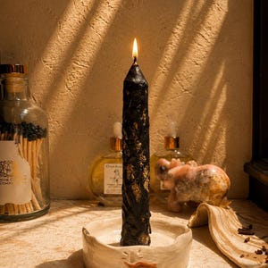 May include: A tall, black taper candle with a textured, dripped wax surface and gold leaf accents, lit and standing upright in a shallow, round, off-white ceramic candle holder featuring a small, hand-painted orange mustache detail.