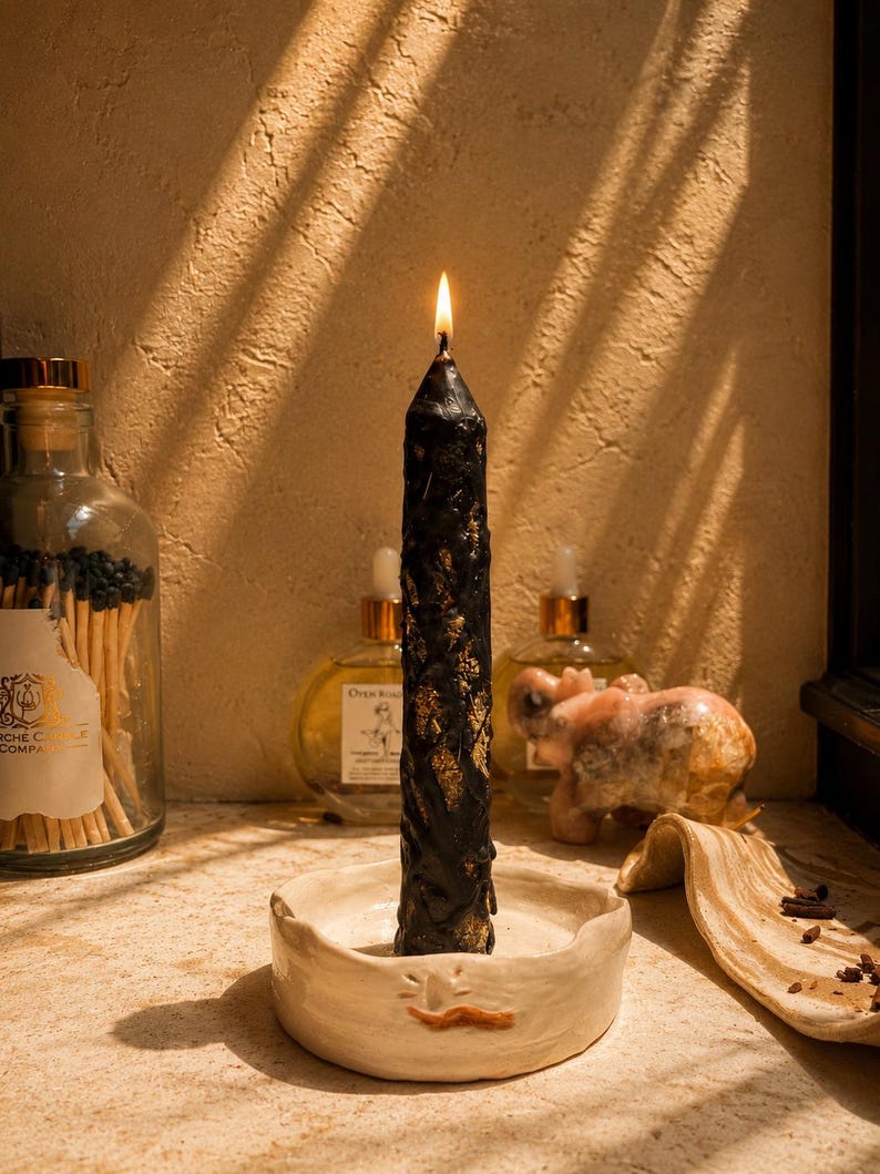 May include: A tall, black taper candle with a textured, dripped wax surface and gold leaf accents, lit and standing upright in a shallow, round, off-white ceramic candle holder featuring a small, hand-painted orange mustache detail.
