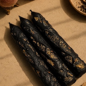 May include: Three cylindrical black candles featuring a textured, raised wax pattern that reveals embedded dried herbs and botanical elements. The candles have a pointed top and a dark, opaque finish, with the organic plant matter visible in the gaps.