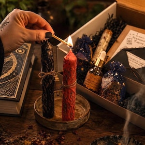May include: A black candle and a red candle stand in a shallow ceramic dish. The red candle is lit with a match. Nearby is an open box containing a glass dropper bottle labeled Love Oil, a blue organza bag with gold stars, and a tarot card set.