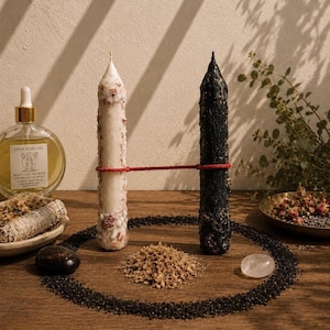 May include: A still life arrangement featuring two tall, textured candles connected by a red cord. One candle is white with pink accents, the other is black. The scene includes a bottle of oil, sage bundles, crystals, and dried herbs, all arranged on a wooden surface.
