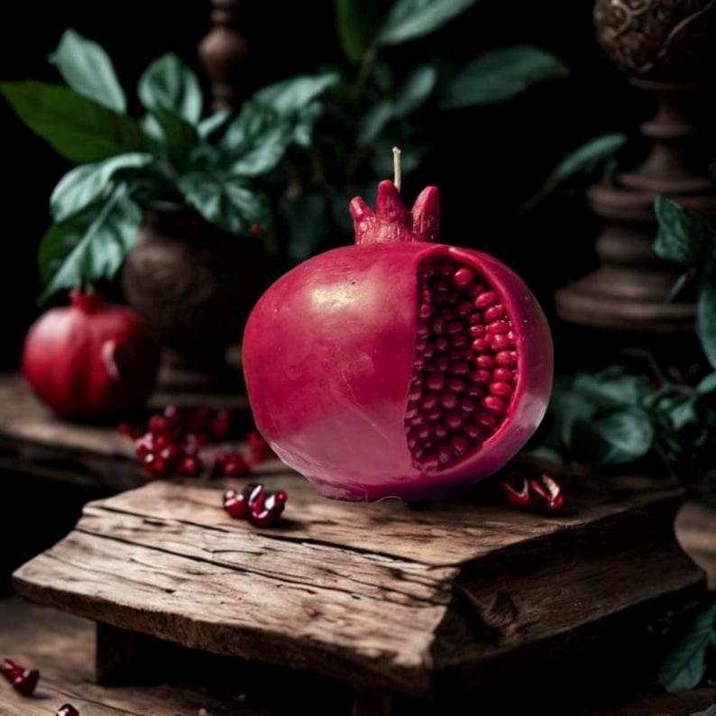 May include: A red pomegranate-shaped candle with a detailed, open section revealing red wax seeds. The candle is sitting on a rustic wooden surface.