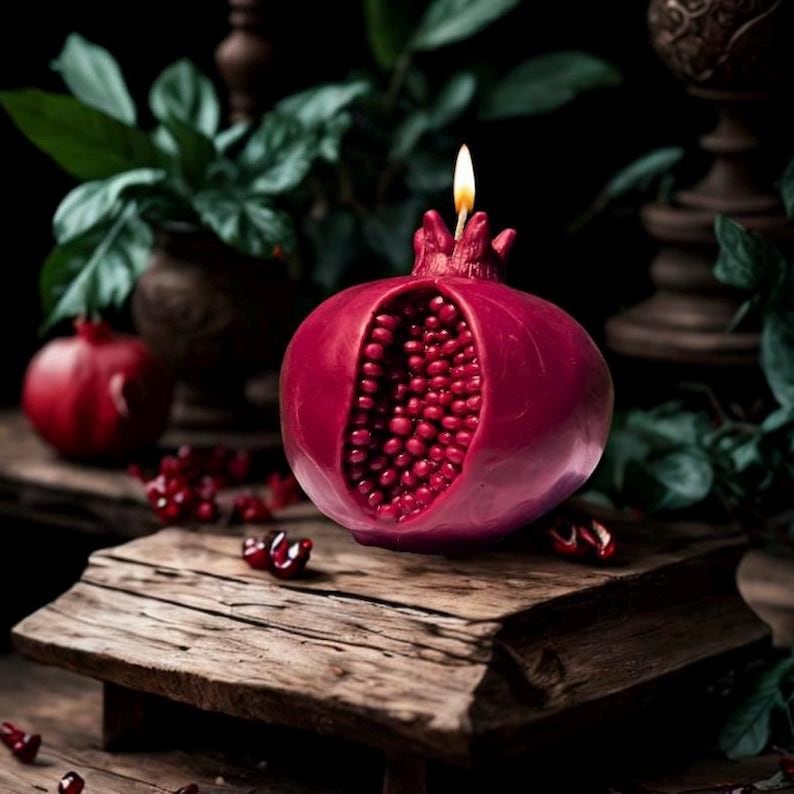 May include: A red pomegranate-shaped candle with a lit wick. The candle is open and shows the red seeds inside. The candle is sitting on a wooden surface.
