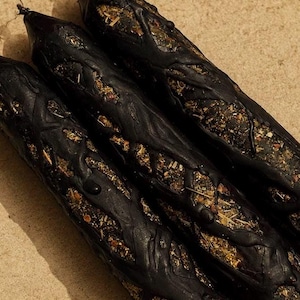 May include: Three cylindrical black candles featuring a textured, raised wax overlay that creates a branch-like pattern. The exposed areas between the black wax reveal a mixture of dried herbs, botanical pieces, and small organic particles.