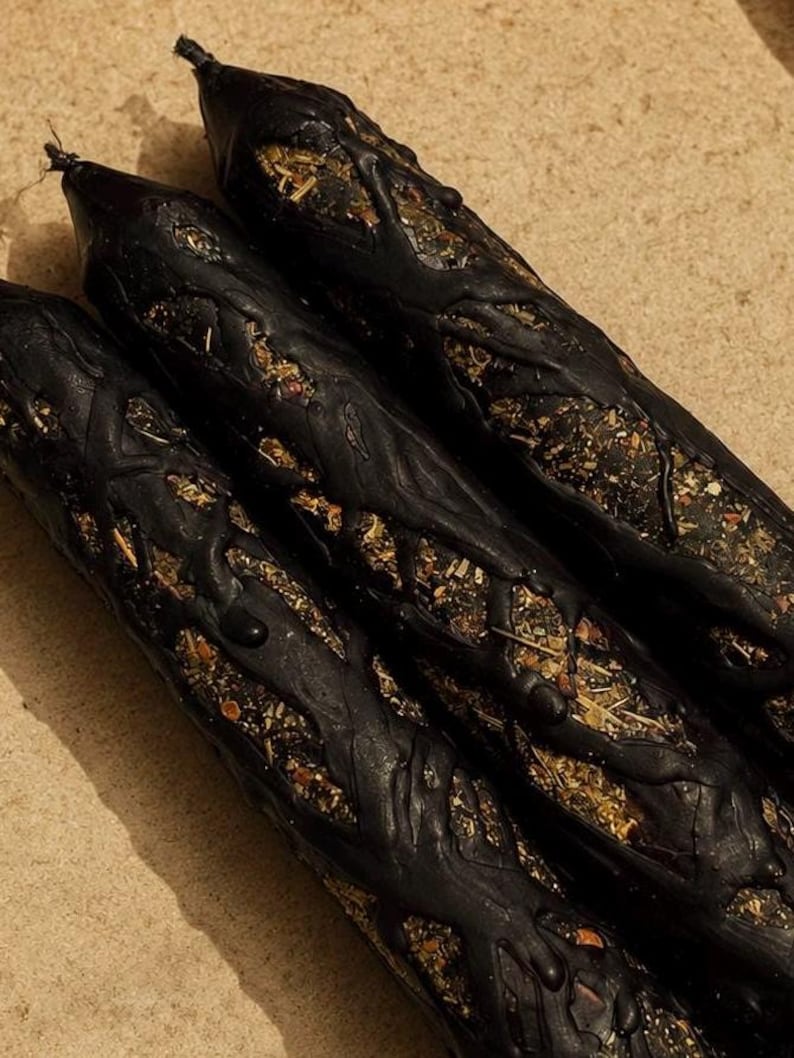 May include: Three cylindrical black candles featuring a textured, raised wax overlay that creates a branch-like pattern. The exposed areas between the black wax reveal a mixture of dried herbs, botanical pieces, and small organic particles.