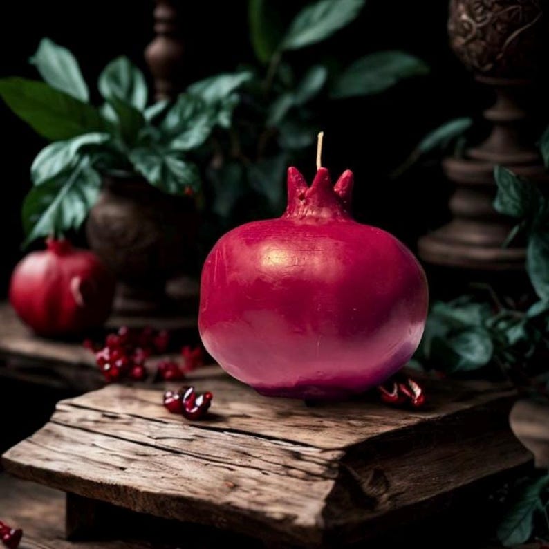 May include: A red pomegranate-shaped candle sits on a rustic wooden surface. The candle is surrounded by small red pomegranate seeds.