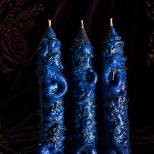May include: Three cylindrical candles featuring a dark blue, textured wax surface with raised, swirling patterns and embedded glitter. Each candle has a pointed top with a visible wick and a deep blue finish that catches the light.