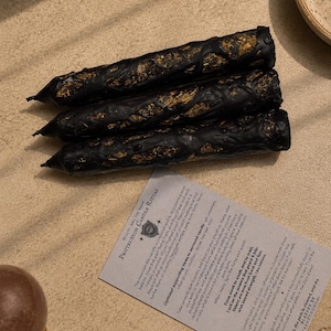 May include: Three black taper candles with textured, uneven surfaces embedded with dried herbs and botanicals. A rectangular instruction card titled Protection Candle Ritual is positioned below the candles, featuring printed text in black ink.