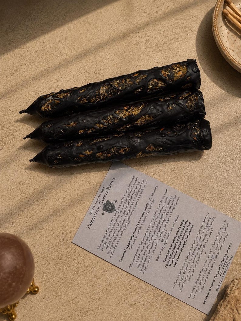 May include: Three black taper candles with textured, uneven surfaces embedded with dried herbs and botanicals. A rectangular instruction card titled Protection Candle Ritual is positioned below the candles, featuring printed text in black ink.