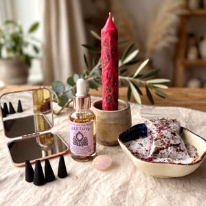 May include: A set featuring a pink textured candle in a stone holder, a glass dropper bottle labeled Self Love, a small rose quartz stone, a folding mirror, four black incense cones, and a bowl containing a plastic bag of floral bath salts.