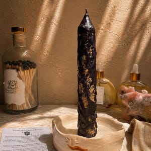 May include: A tall, black, textured pillar candle with visible gold-colored inclusions, standing upright in a shallow, round, off-white ceramic dish. A small paper card titled Protection Candle Ritual is placed next to the candle holder.