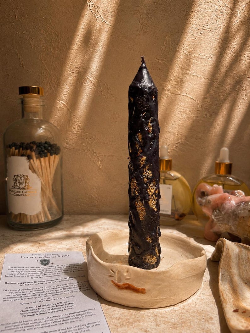 May include: A tall, black, textured pillar candle with visible gold-colored inclusions, standing upright in a shallow, round, off-white ceramic dish. A small paper card titled Protection Candle Ritual is placed next to the candle holder.