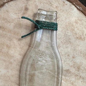 Op de afbeelding: Een doorzichtige glazen fles in de vorm van een wijnfles met een groene touwtje om de hals. De fles heeft een reliëfdesign van twee olijfbomen aan de voorkant.