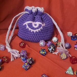 May include: Purple crocheted dice bag with a white eye design. The bag is filled with various colored dice with black numbers.