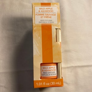 以下が含まれることがあります： 「ワイルドアップル＆アッシュウッド リードディフューザー ディフューザーロゾー 1.01 fl oz (30 mL)」とラベルに書かれたリードディフューザーボトル。ボトルは透明で、表面にテクスチャがあります。ラベルはオレンジと白です。