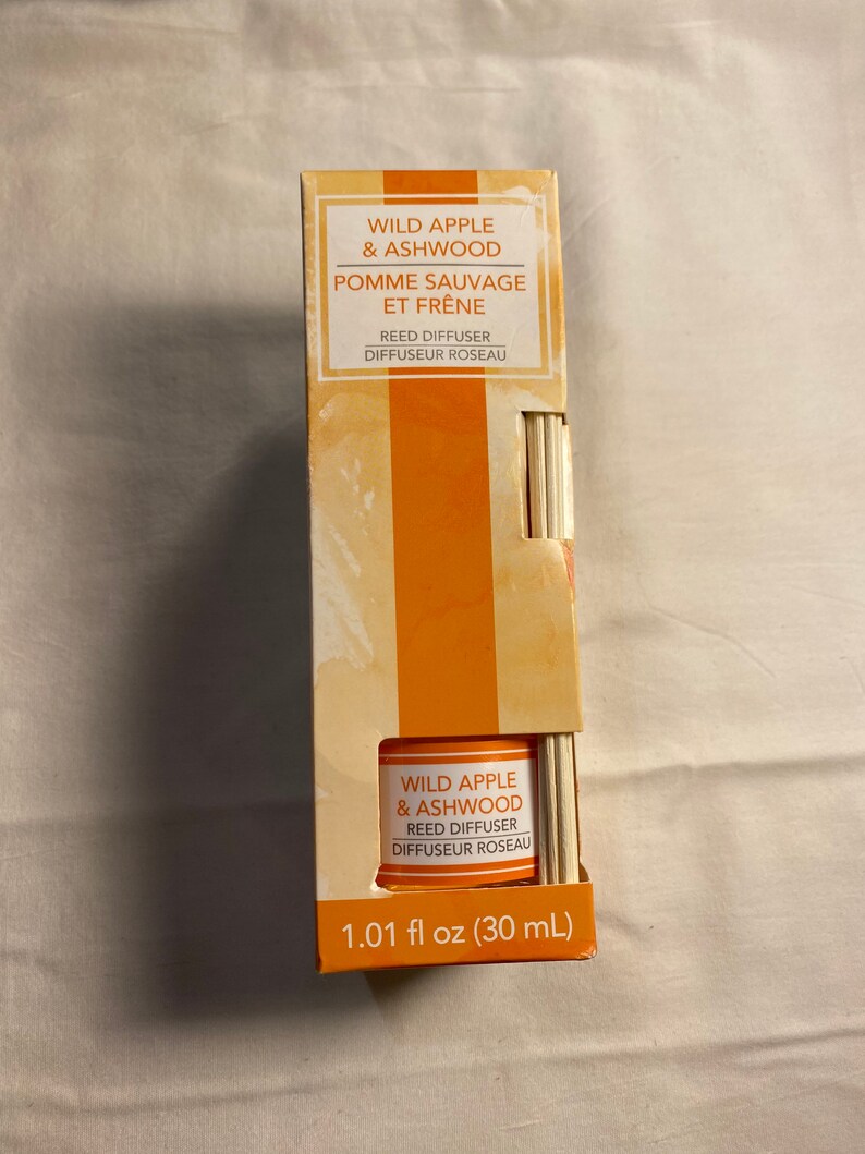 以下が含まれることがあります： 「ワイルドアップル＆アッシュウッド リードディフューザー ディフューザーロゾー 1.01 fl oz (30 mL)」とラベルに書かれたリードディフューザーボトル。ボトルは透明で、表面にテクスチャがあります。ラベルはオレンジと白です。