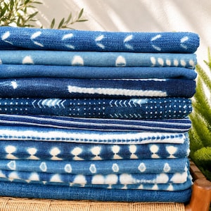 Puede incluir: Una pila de telas dobladas con varios patrones en azul índigo y blanco. Los textiles muestran diversos diseños que incluyen formas geométricas, rayas horizontales, líneas punteadas y motivos abstractos, todos en tonos de azul.