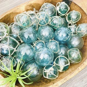 Pode incluir: Uma tigela de madeira cheia de boias de pesca de vidro azul, algumas com rede branca. A tigela está sobre uma superfície de madeira.