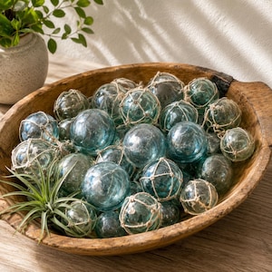 Op de afbeelding: Een verzameling ronde, doorschijnende blauwgroene glazen visdobbers, elk omhuld door een decoratief beige touwnet, geplaatst in een grote, ovale, rustieke houten kom samen met een kleine groene luchtplant.