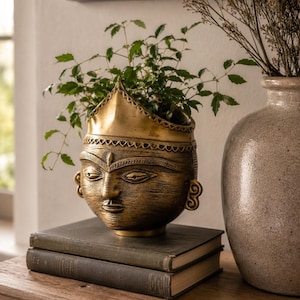 Puede incluir: Una maceta de metal en tono dorado con forma de cabeza humana y borde en forma de corona, con detalles faciales grabados y orejas estilizadas. La maceta contiene una planta verde y descansa sobre dos libros de tapa dura de color oscuro.
