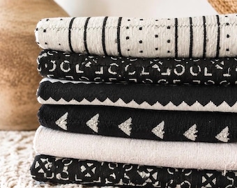 May include: A stack of six folded textiles featuring various black and white patterns. The designs include vertical stripes with dots, repeating geometric shapes, a zigzag border, small triangles, a solid cream layer, and a complex cross-like motif.