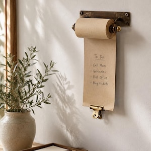 Può includere: Un portarotolo da parete in metallo con finitura bronzo scuro, contenente un rotolo di carta kraft marrone chiaro. La carta mostra una lista di cose da fare scritta a mano: Call Mom, Groceries, Post Office, Buy tickets, fissata da una clip.